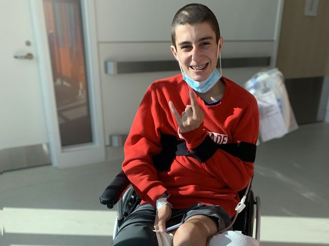 A shaven-headed teenager, smiling, sitting in a wheelchair, with a thick bandage on his left leg. 