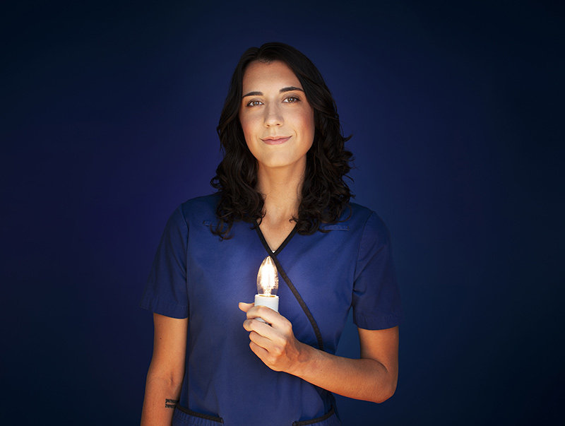 Sandrine, infirmière, porte un uniforme bleu et tient fièrement une ampoule allumée dans sa main gauche.