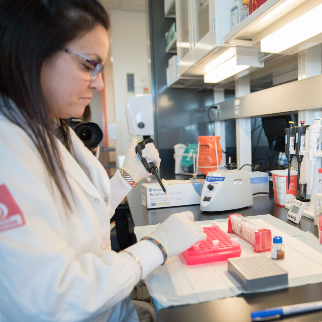 Une chercheuse portant une blouse blanche et des gants de protection manipule un échantillon dans un laboratoire. 