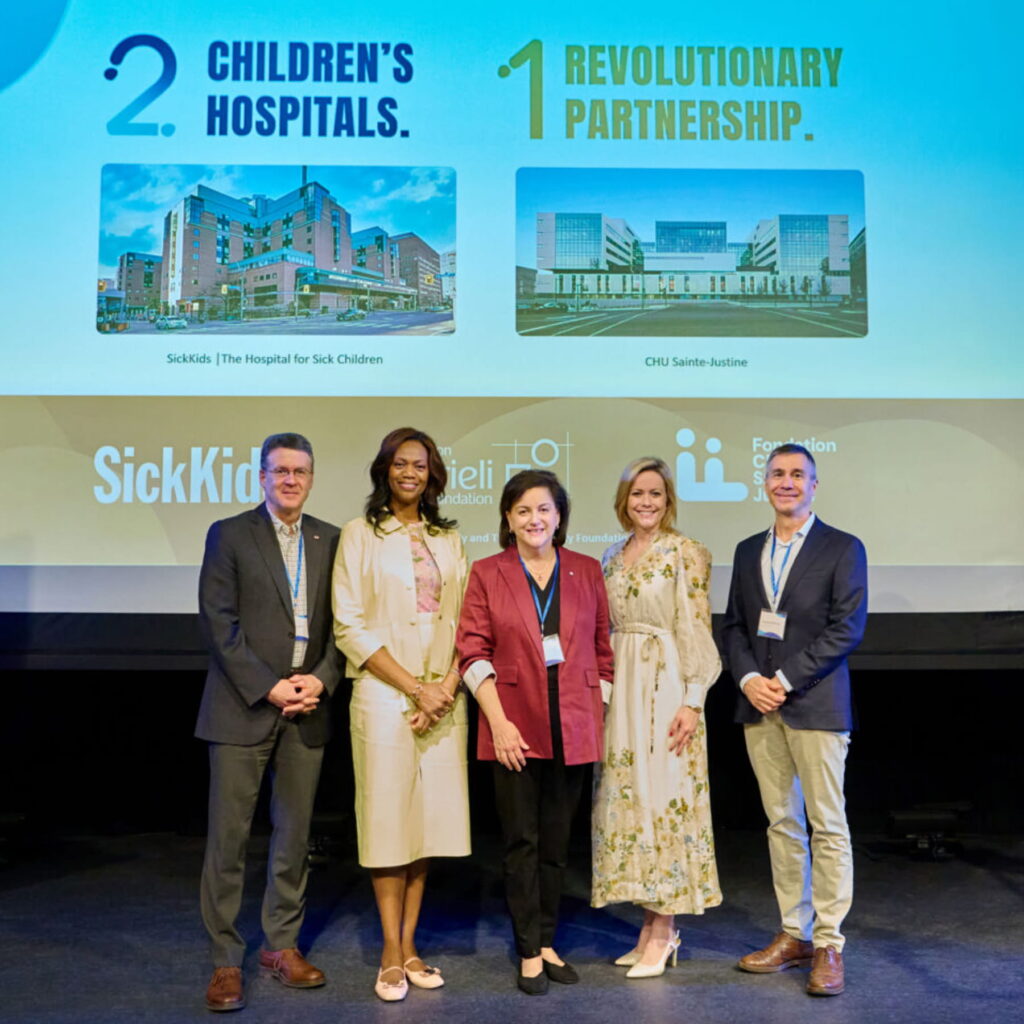 Standing on a stage, in front of a screen showing the two partner hospitals, five people smile at the camera.