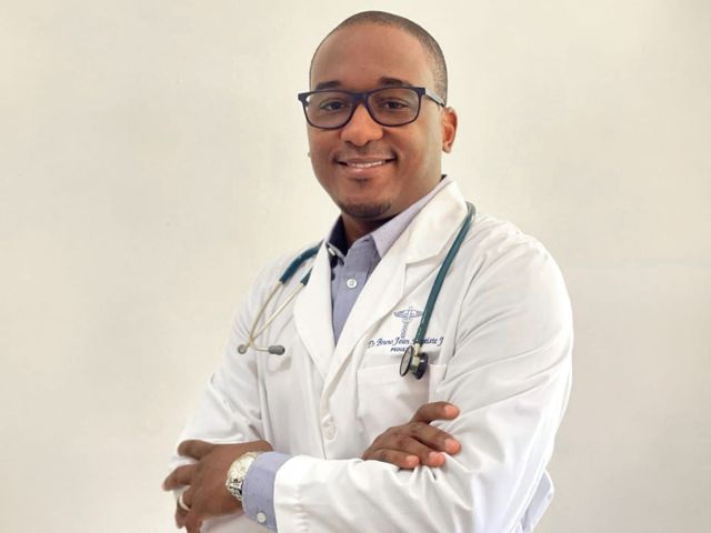 Un médecin en sarrau blanc, stéthoscope au cou, sourit en regardant la caméra.