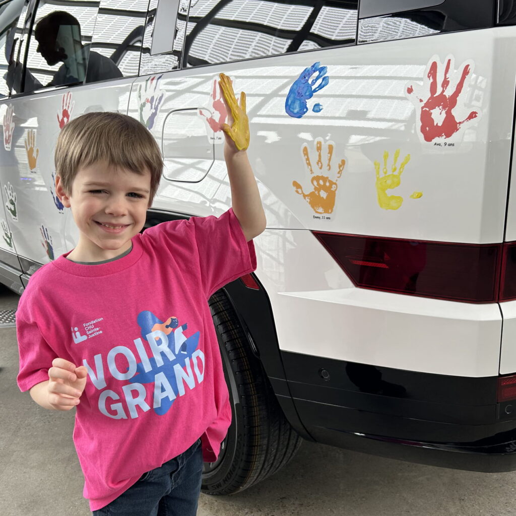Devant un véhicule blanc couvert de petites empreintes de mains colorées, un enfant en t-shirt rose « Voir Grand » sourit en levant sa main peinte en jaune.