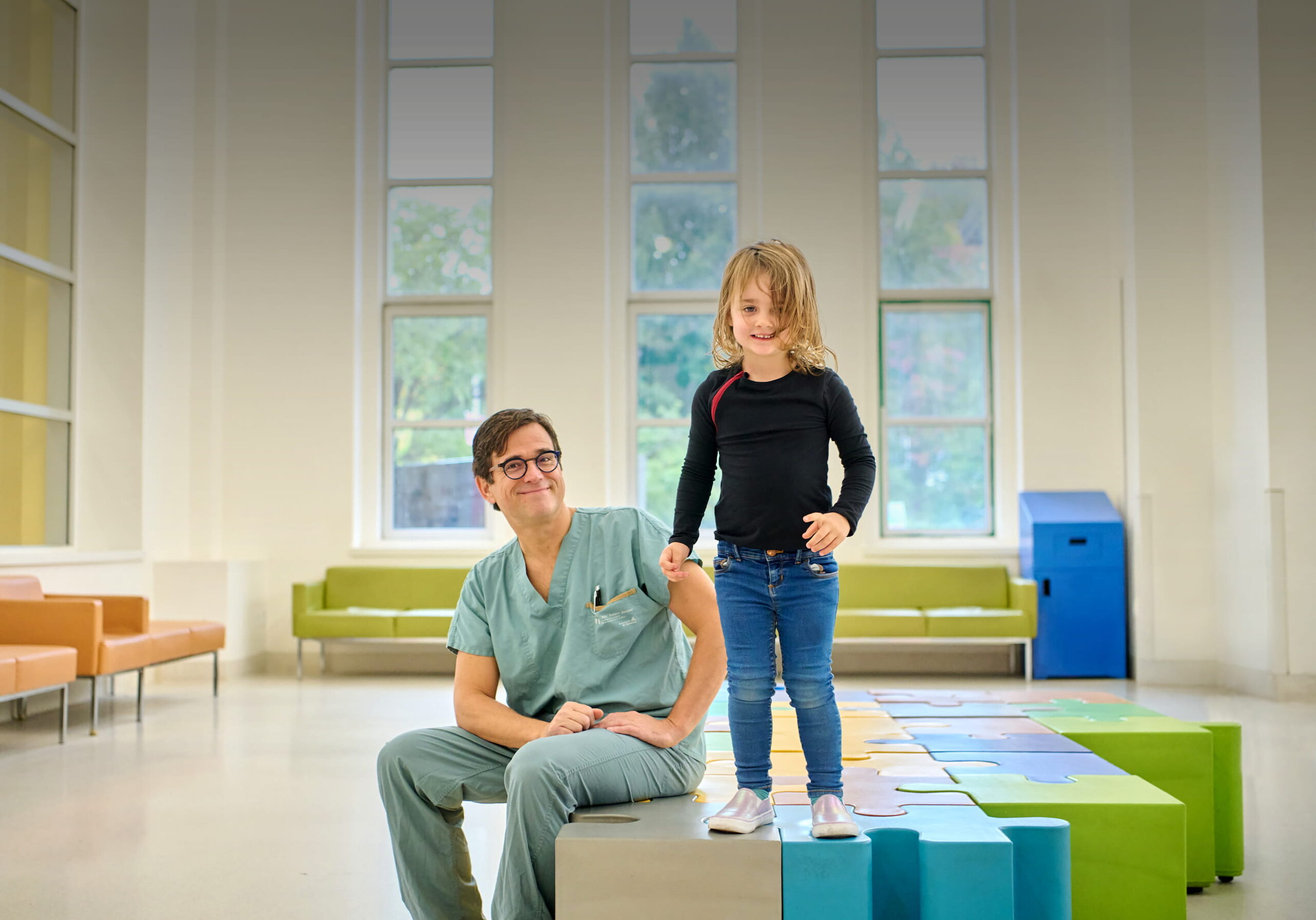 Dans une salle lumineuse aux grandes fenêtres, le Dr Nelson Piché, en uniforme vert, est assis sur un banc coloré à côté de la petite Françoise, debout et souriante.
