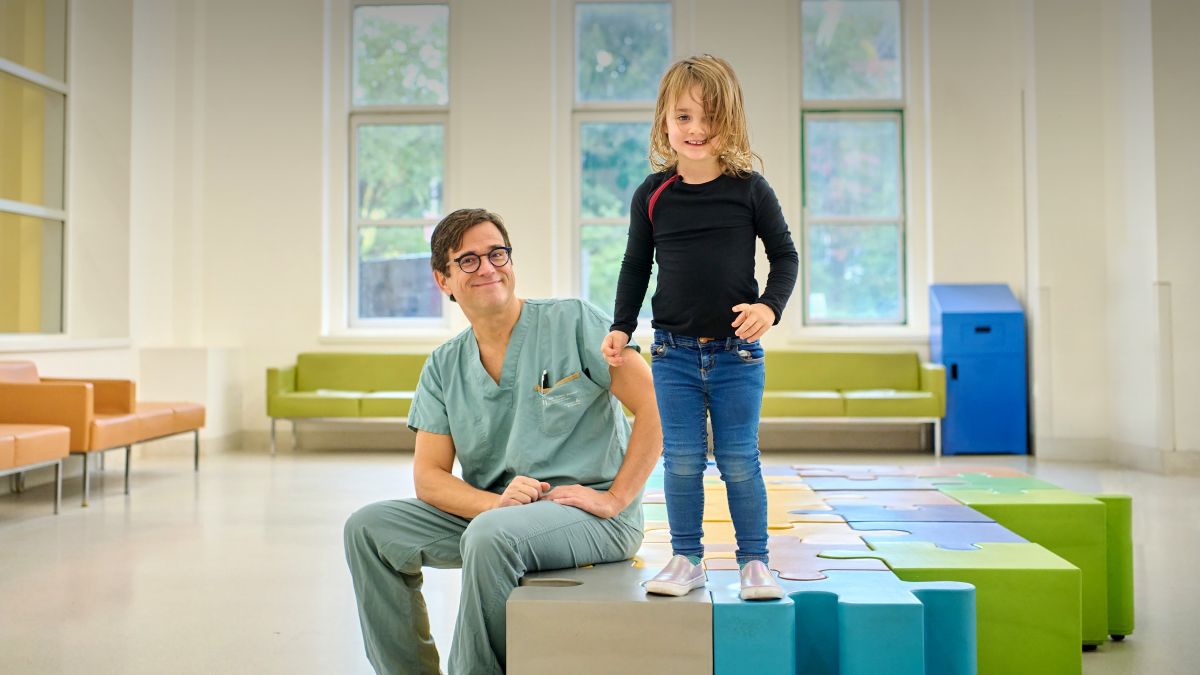 Dans une salle lumineuse aux grandes fenêtres, le Dr Nelson Piché, en uniforme vert, est assis sur un banc coloré à côté de la petite Françoise, debout et souriante.