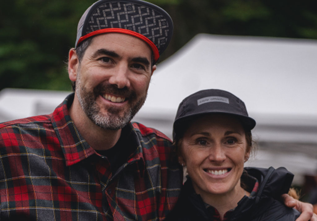 Deux personnes souriantes portant des casquettes, devant des tentes lors d&rsquo;un &eacute;v&eacute;nement.