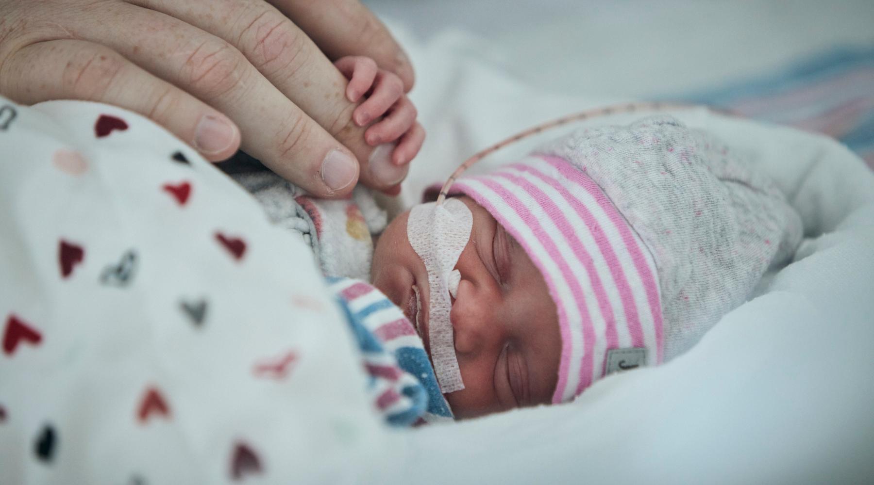 Elyrose, un b&eacute;b&eacute; &agrave; l'unit&eacute; de n&eacute;onatalogie du CHU Sainte-Justine, est couch&eacute;e, les yeux ferm&eacute;s, et tient le doigt de son papa avec sa minuscule main.
